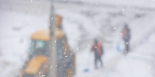 在冬天的暴风雪中清扫街道。工人们拿着铁锹和带着铲斗的拖拉机在离焦的道路上清理积雪。选择性聚焦，浅景深。大雪