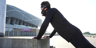 侧面的观点，有吸引力的自信活跃的大胡子男人面具在黑色运动服，做俯卧撑在一个具体的支持在现代体育场