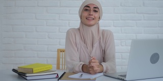 一位年轻的穆斯林妇女戴着希贾布坐在桌旁微笑，她很高兴，因为她可以接受教育。桌上有笔记本电脑和书。穆斯林妇女远程教育的概念