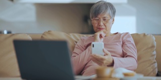 亚洲中国高级妇女使用智能手机在线购物在她的公寓在城市的客厅