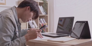 一位亚洲设计师用他的智能手机交流。完成项目和论文。在家里用笔记本电脑在桌子上工作。