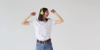 一个穿着白色t恤的年轻美女戴着耳机听音乐，跳舞，闲逛，愉快地做着手势，在白色的背景下享受假期。