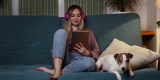 一个女孩躺在沙发上，一只狗趴在她的笔记本电脑上，戴着耳机听音乐。自由职业和通过互联网交流