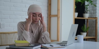不堪重负的穆斯林学生，戴着头巾的阿拉伯年轻女子，思考着，压力在远程学习。一名女子因训练过度而摩擦太阳穴，头痛