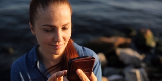 年轻漂亮的红发白人女子，手拿手机，坐在海边的一块大石头上，在社交网络上交流