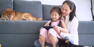母女俩和柴犬一起坐在沙发旁边看数码平板电脑。快乐的母女在家使用平板电脑。