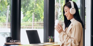 一个亚洲女人坐在咖啡店里，拿着笔记本电脑喝咖啡，然后看向别处。亚洲女性放松在café。
