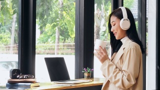 一个亚洲女人坐在咖啡店里，拿着笔记本电脑喝咖啡，然后看向别处。亚洲女性放松在café。视频素材模板下载