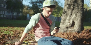 英俊的年轻人在秋季公园坐在滑板上发短信的手机
