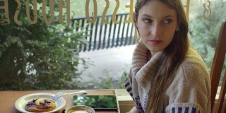 一个年轻的女人坐在咖啡馆的一张桌子旁，一边喝着咖啡，一边看着窗外