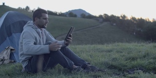 一个年轻人坐在帐篷附近的户外山上，在日出日落时使用平板电脑，然后走开——慢镜头高清视频