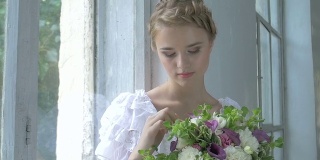 美丽的女孩，穿着复古的白色连衣裙，坐在窗台上触摸鲜花，微笑，慢镜头