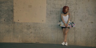 有纹身的红发女人和他的滑板站在一堵水泥墙里