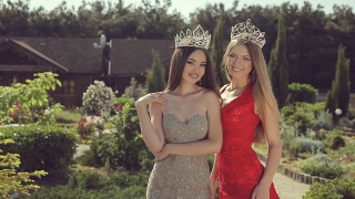 两个美丽的年轻女孩穿着晚礼服，戴着皇冠，微笑着站在一个绿色的公园里视频素材模板下载