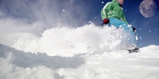 一名滑雪者在蓝天上有月亮的情况下从雪坡上滑下来的有力而充满活力的动态镜头。先是实时速度视频，然后减速来展示猛烈的雪暴。