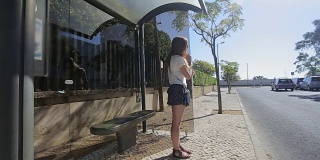 年轻女孩在公交车站等待座位与智能手机在蓝色裙子和太阳镜，夏季，和交流
