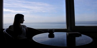 长剪影的年轻美丽的女孩坐在咖啡馆与大西洋的全景。早上喝一杯咖啡，喝茶，看书，喝酒，思考