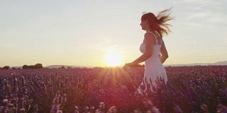 慢镜头:在金色的夏日落日中，身着白裙的快乐年轻女子奔跑在美丽的紫色薰衣草田中