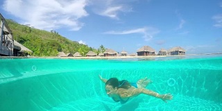 慢镜头:女子在水下游泳池游泳，背景是水上别墅