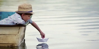 戴着柳条帽的男孩坐在船上，坐着他的纸船去旅行