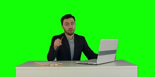 快乐的商人坐在办公室里拿着笔记本电脑对着绿色屏幕的摄像头说