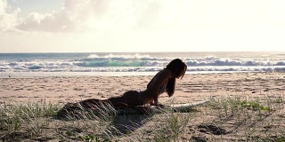 健康女孩伸展在冲浪板上在日出的海滩上。幻灯片。缓慢的运动。