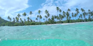 4K慢镜头半水下:令人惊叹的水晶清澈的海洋泻湖在异国情调的白色沙滩与高大茂盛的棕榈树生长在热带岛屿在阳光明媚的夏天