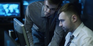 两名IT程序员正在一间满是显示屏的黑暗办公室里用电脑工作。