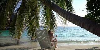 年轻苗条的女人享受假期在一个热带海滩的太阳躺椅上。