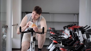 胖子转动着健身自行车的踏板，手里拿着一杯饮料和一片披萨视频素材模板下载