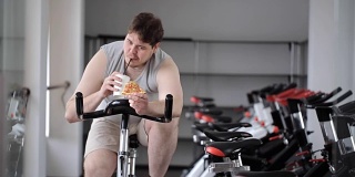 胖子转动着健身自行车的踏板，手里拿着一杯饮料和一片披萨