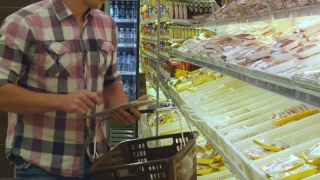 人们推着购物车在超市购买冷藏食品，用平板电脑查看购物清单。一个人走到商店的冰箱前，拿起商品放进篮子里视频素材模板下载
