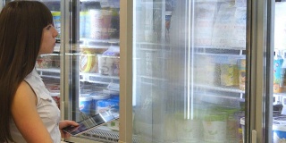 年轻女子在超市的冷藏区打开玻璃门购买乳制品或冷藏食品。女孩在商店从冰箱里拿东西，用平板电脑检查购物清单