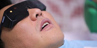 在紫外线辐射下戴墨镜的亚洲人男性病人在牙科椅上张开他的嘴