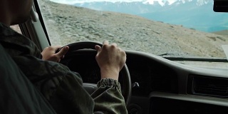 从SUV汽车内部的部分内饰和镜子与司机男性的手在方向盘上危险笔直的山路在阳光明媚的日子与雪山的背景