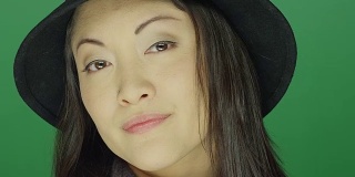 美丽的年轻亚洲女人戴着一顶帽子，看起来严肃，在一个绿色屏幕的工作室背景