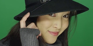 美丽的年轻亚洲女人在一顶帽子微笑和调情，在一个绿色屏幕工作室的背景