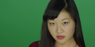 一名年轻的亚洲女子转过身来，盯着录音室背景的绿色屏幕上的摄像机