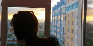 年轻女子拉开窗帘，在美丽的夕阳下从窗口俯视城市