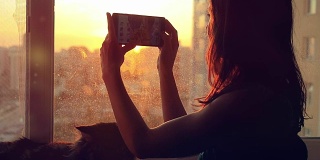 戴着耳机的年轻女子与她可爱的缅因猫在窗口拍摄了日落时模糊的城市背景的慢动作