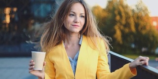 年轻漂亮的女孩穿着黄色夹克在办公室附近停留，微笑着在阳台上喝着咖啡。商务中心微笑着喝茶的商务女性。早晨明亮的阳光