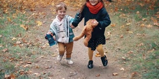可爱的兄妹小男孩和小女孩手牵着手奔跑着，带着他们的玩具穿过秋天的公园慢莫胡同