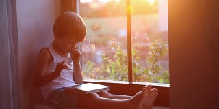 可爱的小男孩，玩着平板电脑，坐在窗罩上，夕阳落在窗后