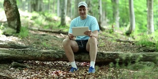 一名手持平板电脑的男子坐在森林里倒下的一棵树上