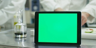 在一间商业厨房的桌子上放着一台绿色屏幕的平板电脑，背景是厨师正在准备食物。