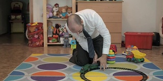 成年男性商人玩玩具铁路，喜欢坐在婴儿室的地板上。