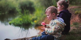 侧面是两个可爱的小孩，一个男孩和一个女孩，坐在河边吃西瓜