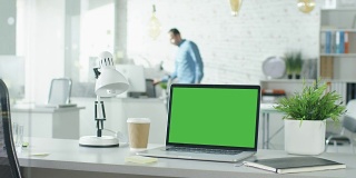 绿色屏幕的笔记本电脑站在桌子上，没有人在上面工作。背景里，一个男人走进来，坐在他的工作场所开始工作。