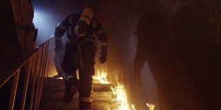 两名强壮的消防员在燃烧的大楼里上楼。楼梯被明火燃烧。在缓慢的运动。