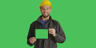 中景微笑工作与安全帽上持有水平平板电脑与绿色屏幕。背景中的绿色屏幕。